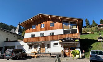 A wooden building with a business called "KäseStadl". In front of the house are several cars, and green hills can be seen in the background. | © Käsestadl Kleinwalsertal | Nadine Paul