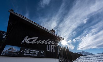 A building with the inscription "Kessler" and a sunburst in the background. The sky is blue with some clouds and the surroundings look alpine. | © Sport Kessler GmbH