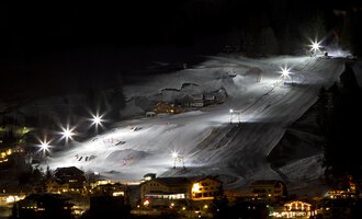 A snowy ski landscape at night with illuminated slopes. In the background, small huts and a clear sky can be seen. | © Sport Kessler GmbH