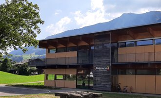 A modern building with large windows and wooden cladding. In the foreground, there is a stone table on a green meadow with mountains in the background. | © Kleinwalsertal Tourismus | Veronika Senn