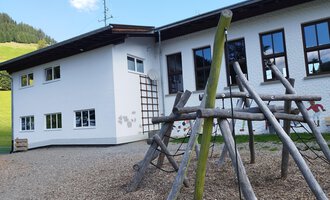 A modern building with large windows and a wooden playground in the foreground. The playground is made of natural materials and offers various play opportunities. | © Kleinwalsertal Tourismus | N. Lughammer