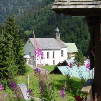 A picturesque landscape with a church and colorful flowers in the foreground. Tall trees and gentle hills surround the scene. | © Kleinwalsertal Tourismus | Rolf Köberle