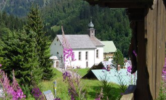 A picturesque landscape with a church and colorful flowers in the foreground. Tall trees and gentle hills surround the scene. | © Kleinwalsertal Tourismus | Rolf Köberle