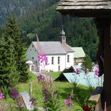 A picturesque landscape with a church and colorful flowers in the foreground. Tall trees and gentle hills surround the scene. | © Kleinwalsertal Tourismus | Rolf Köberle