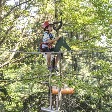Ein Junge sitzt in einem Hochseilgarten auf einem Seil. Die Umgebung ist von Bäumen umgeben, und er trägt einen Helm und Sicherheitsausrüstung. | © OBERSTDORF · KLEINWALSERTAL BERGBAHNEN