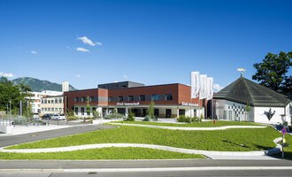 A modern building with a curved lawn area and a clear blue sky. In the background, trees and mountains are visible. | © Klinikverbund Allgäu
