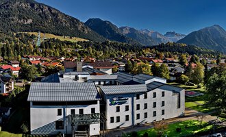 A modern building surrounded by mountains and trees. The sky is clear and the surroundings feel calm and inviting. | © Klinikverbund Allgäu