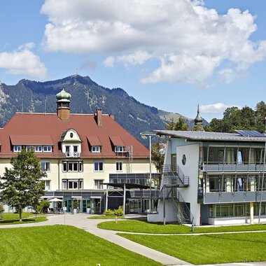 A charming hotel building with a red roof and modern architecture. In the background, mountains and a blue sky can be seen. | © Klinikverbund Allgäu