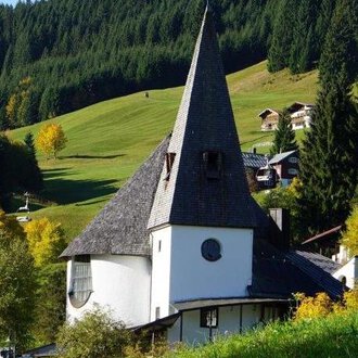 A charming, modern building with a tall pointed roof stands amidst a green landscape. Surrounded by trees and gentle hills, the scene radiates tranquility and beauty. | © Ev. Kirche Kleinwalsertal