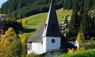 A charming, modern building with a tall pointed roof stands amidst a green landscape. Surrounded by trees and gentle hills, the scene radiates tranquility and beauty. | © Ev. Kirche Kleinwalsertal