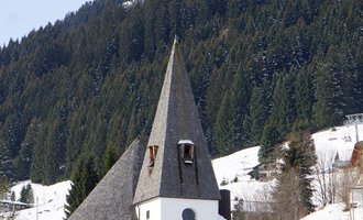 A picturesque church with a pointed tower stands in the snow. Forested mountains rise in the background. | © Ev. Kirche Kleinwalsertal