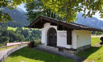 A small, white building with a gabled roof stands in a green landscape. In the background, gentle hills and trees can be seen. | © Kleinwalsertal Tourismus | Nicole Lughammer