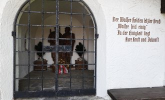 A historical entrance with a sturdy wrought-iron gate. Inside, there are statues adorned with flowers. | © Kleinwalsertal Tourismus | Nicole Lughammer