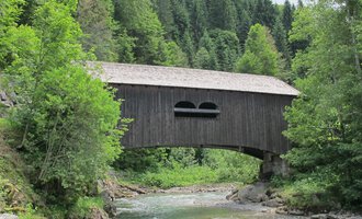 An old wooden bridge over a clear river, surrounded by green trees. In the background, wooded hills rise up. | © Stefan Heim
