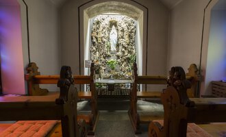 A quiet chapel with wooden benches and a statue in the niche. The walls are softly illuminated, creating a peaceful atmosphere. | © Kleinwalsertal Tourismus | Oliver Farys