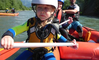 A child in a rowing boat is wearing a helmet and a life jacket. In the background, other boaters can be seen on a river. | © MAP-Erlebnis