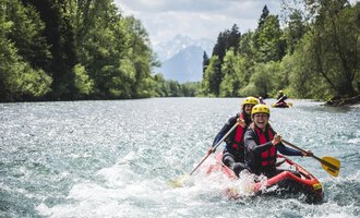 A group of people is paddling in a raft on a clear river. In the background, trees and mountains are visible. | © MAP-Erlebnis