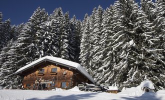 A cozy wooden house in the snow, surrounded by tall, snow-covered fir trees. The clear blue sky creates a picturesque winter landscape. | © Kleinwalsertal Tourismus | Oliver Farys