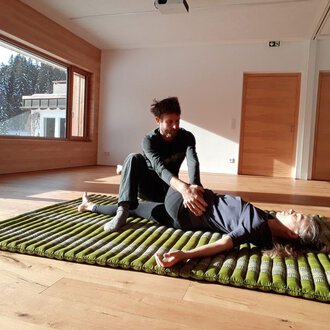 A person is lying on a mat while another person gently treats them. The room has large windows overlooking trees and plenty of daylight. | © Alexander Mayr