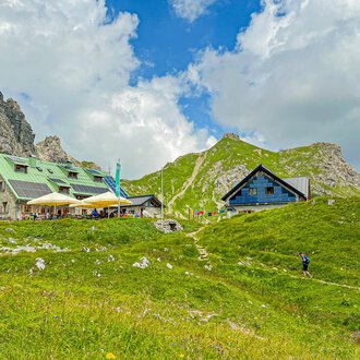 A picturesque mountain landscape with cozy cottages and green meadows. In the background, impressive rocks and a blue sky with clouds can be seen. | © Mindelheimer Hütte | Wolfgang Stache