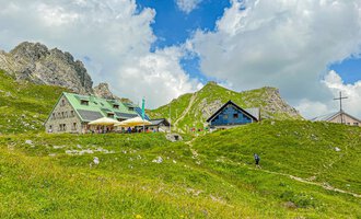 A picturesque mountain landscape with cozy cottages and green meadows. In the background, impressive rocks and a blue sky with clouds can be seen. | © Mindelheimer Hütte | Wolfgang Stache