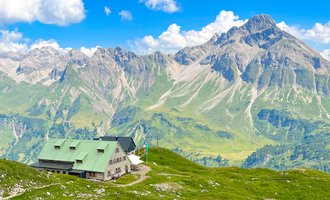 A mountain cabin is picturesque on a green meadow. In the background, impressive mountains rise under a blue sky with some clouds. | © Mindelheimer Hütte | Wolfgang Stache