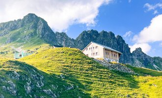 A typical mountain house stands on a green meadow with majestic mountains in the background. The sky is blue and partly cloudy. | © Mindelheimer Hütte | Wolfgang Stache