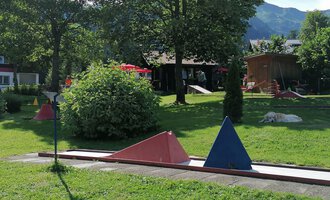 A mini golf course with colorful obstacles in a green environment. In the background, trees and part of a building can be seen. | © Kleinwalsertal Tourismus | N. Lughammer