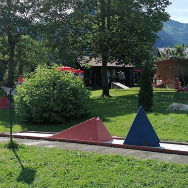 A mini golf course with colorful obstacles in a green environment. In the background, trees and part of a building can be seen. | © Kleinwalsertal Tourismus | N. Lughammer