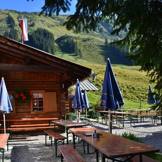 A cozy cabin in the mountains with tables and benches outdoors. In the background, green meadows and mountains can be seen. | © Kleinwalsertal Tourismus