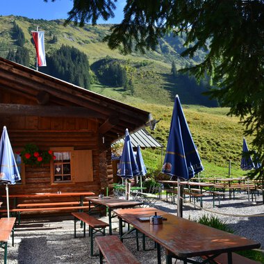 A cozy cabin in the mountains with tables and benches outdoors. In the background, green meadows and mountains can be seen. | © Kleinwalsertal Tourismus