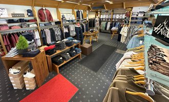 A fashionable clothing store with a variety of shirts and sweaters. The shelves are neatly organized, and the floor is covered with a red carpet as well as a patterned rug. | © Moosbrugger Mode e.U.
