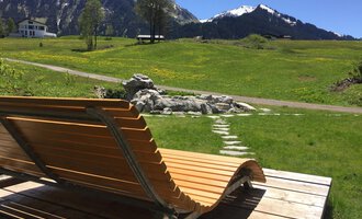 A wooden lounge chair is on a terrace overlooking green meadows and snow-capped mountains in the background. The sky is clear and the landscape looks idyllic. | © Kleinwalsertal Tourismus | Sarina Berchtold