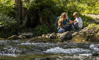 A mother and her son are sitting by a clear stream in nature. They are having fun and enjoying their time together. | © Kleinwalsertal Tourismus | Frank Drechsel