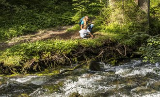 Two people are sitting by the bank of a babbling brook, surrounded by green nature. The sun is shining through the trees, and there is a calm atmosphere. | © Kleinwalsertal Tourismus | Frank Drechsel