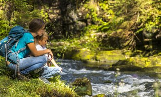 A woman sits with a child by the bank of a babbling brook. Both enjoy nature and the peaceful surroundings. | © Kleinwalsertal Tourismus | Frank Drechsel