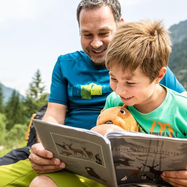 A father and his son are sitting outside and reading a book together. Both are smiling and the boy is holding a stuffed animal in his hand. | © Kleinwalsertal Tourismus | Bastian Morell
