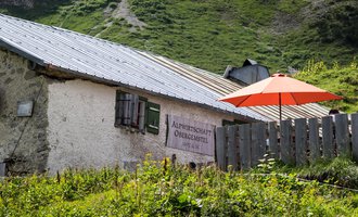 A charming alpine hut with a red sun umbrella in the greenery. In the background, gentle hills and a mountain landscape can be seen. | © Kleinwalsertal Tourismus | Andre Tappe