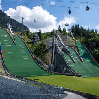 Zwei große Sprungschanzen umgeben von grünen Wiesen und Bergen. Im Hintergrund hängen Seilbahnen unter einem blauen Himmel. | © Skiclub Oberstdorf Veranstaltungs GmbH