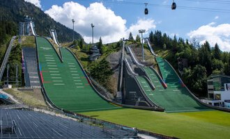 Zwei große Sprungschanzen umgeben von grünen Wiesen und Bergen. Im Hintergrund hängen Seilbahnen unter einem blauen Himmel. | © Skiclub Oberstdorf Veranstaltungs GmbH