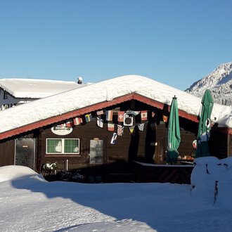A cozy cabin in the snow, surrounded by a wintry landscape. The sky is clear and the mountains are clearly visible.