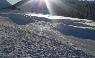A snowy landscape under a clear sky. The sun shines brightly behind the mountains.