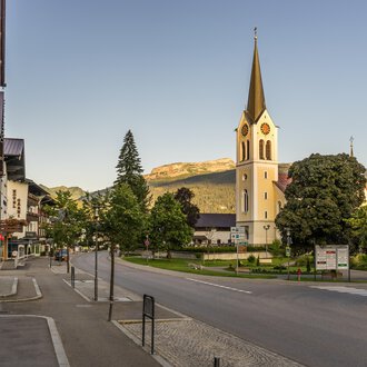 A quiet street in a charming village with a tall church in the background. Surrounded by trees and mountains, the scene radiates tranquility. | © Kleinwalsertal Tourismus | Steffen Berschin