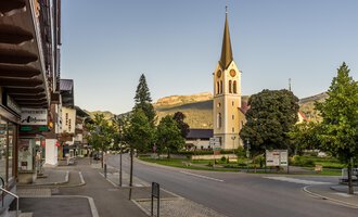 A quiet street in a charming village with a tall church in the background. Surrounded by trees and mountains, the scene radiates tranquility. | © Kleinwalsertal Tourismus | Steffen Berschin