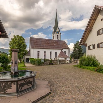 A picturesque village square with a fountain in the foreground and traditional houses. In the background, there is a church with a tall tower and a cloudy sky. | © Kleinwalsertal Tourismus | Steffen Berschin
