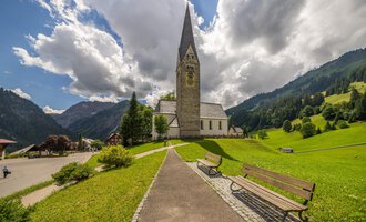 A picturesque church with a pointed tower stands in a green landscape. In the background, gentle hills and clouds in the sky can be seen. | © Kleinwalsertal Tourismus | Steffen Berschin