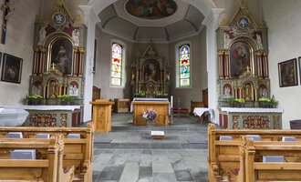 A beautiful church interior with rows of wooden benches and colorful windows. The altar is centrally located and surrounded by artistic decorations. | © Kleinwalsertal Tourismus | Oliver Farys