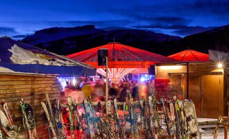 Eine Skihütte bei Nacht mit rot beleuchteten Schirmen und vielen Skiern im Vordergrund. Im Hintergrund sind verschwommene Menschen zu sehen, die eine gesellige Atmosphäre genießen. | © Plattform Kleinwalsertal | Michael Monschau