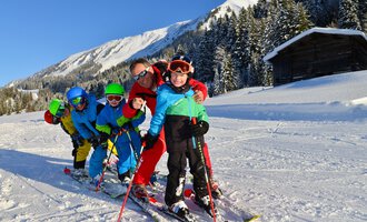 A group of children and an adult skiing in the snowy mountain landscape. They are wearing colorful ski clothes and smiling at the camera. | © Andy Herr | Werbewind GmbH