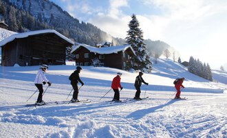 A group of skiers is skiing on a snowy slope. In the background, wooden huts and snow-covered mountains can be seen. | © Andy Herr | Werbewind GmbH